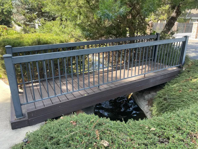 Pedestrian bridge with metal railing and trex decking, surrounded by manicured shrubs