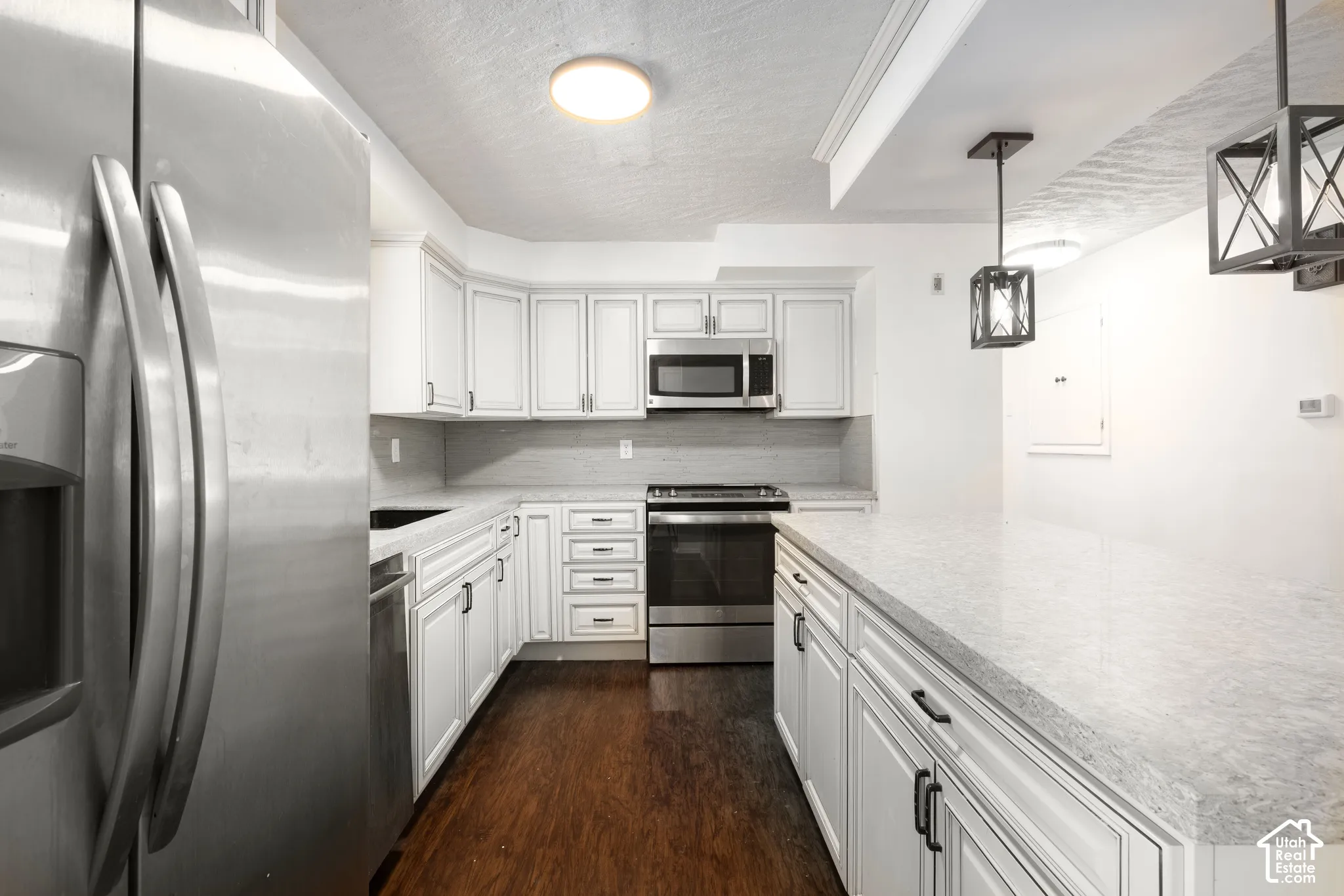 White shaker cabinetry with black hardware, stainless steel appliances, light-toned countertops, wood-finish flooring, and two pendant light fixtures
