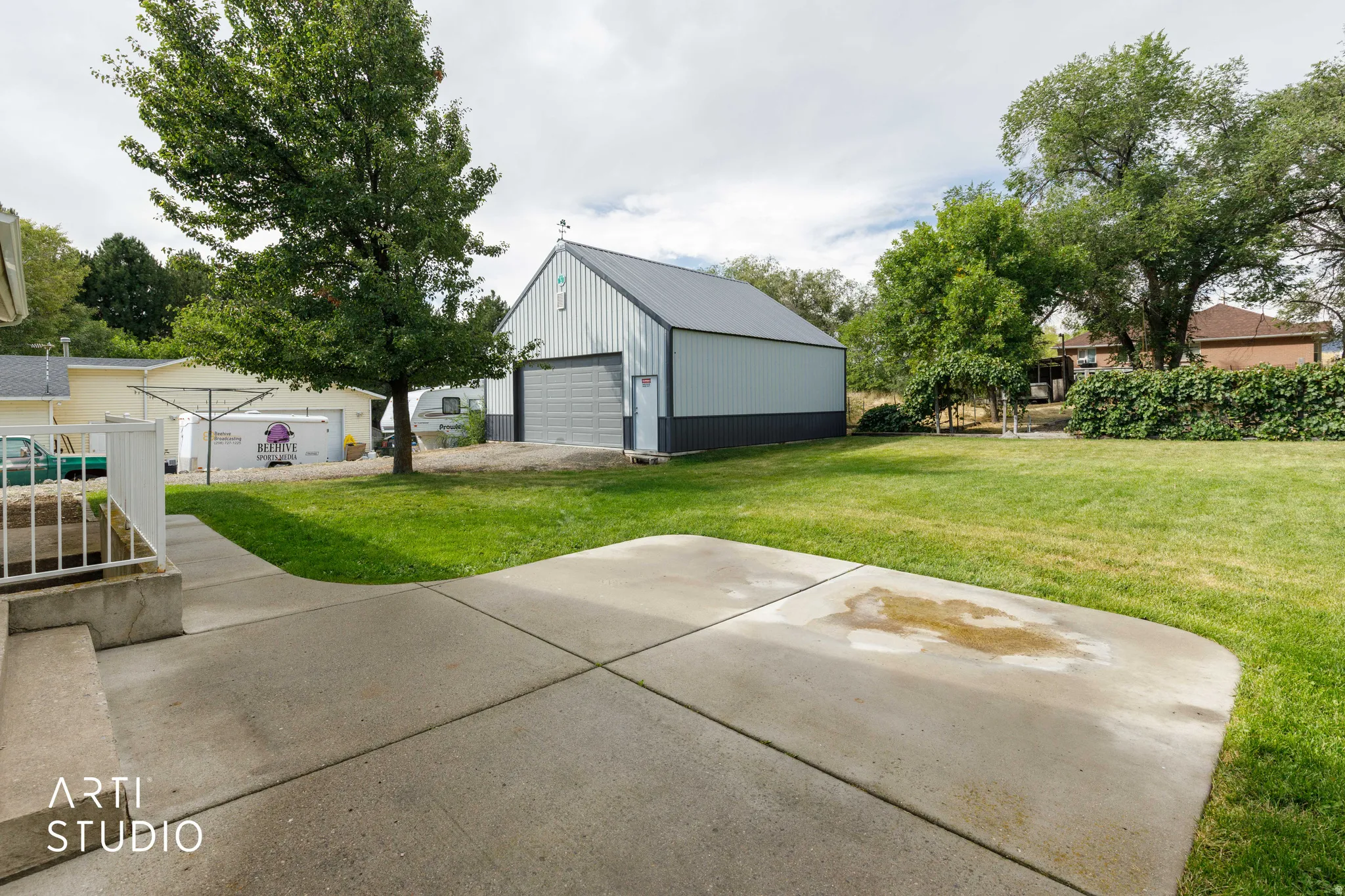 Expansive yard with a large concrete patio, mature trees, and a detached metal-sided building featuring a roll-up door