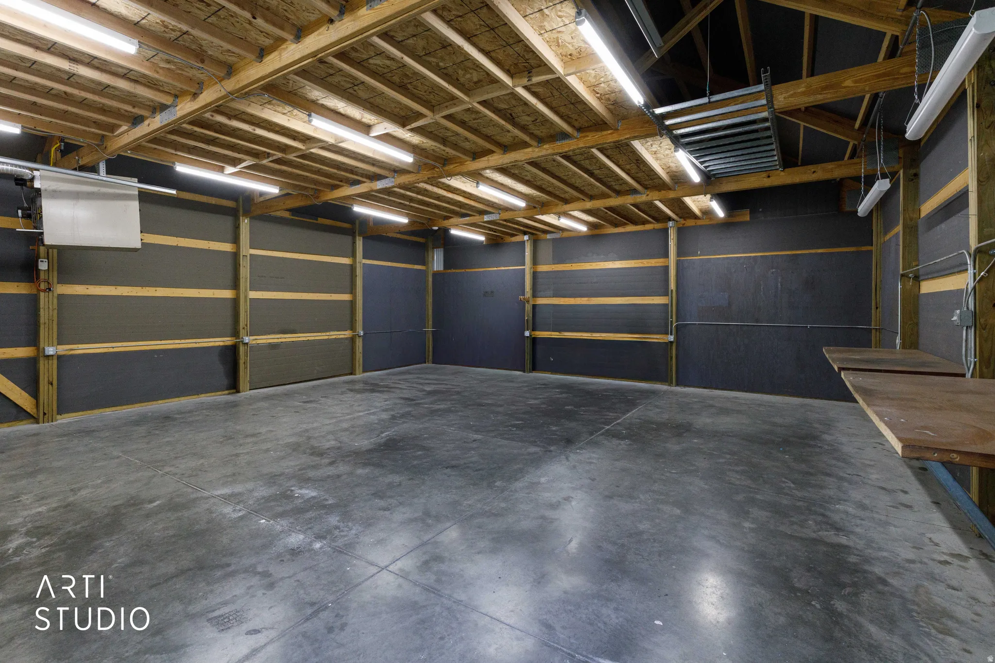 Spacious shop interior with exposed wood beam ceiling, multiple fluorescent light fixtures, concrete flooring, and built-in wooden work surfaces