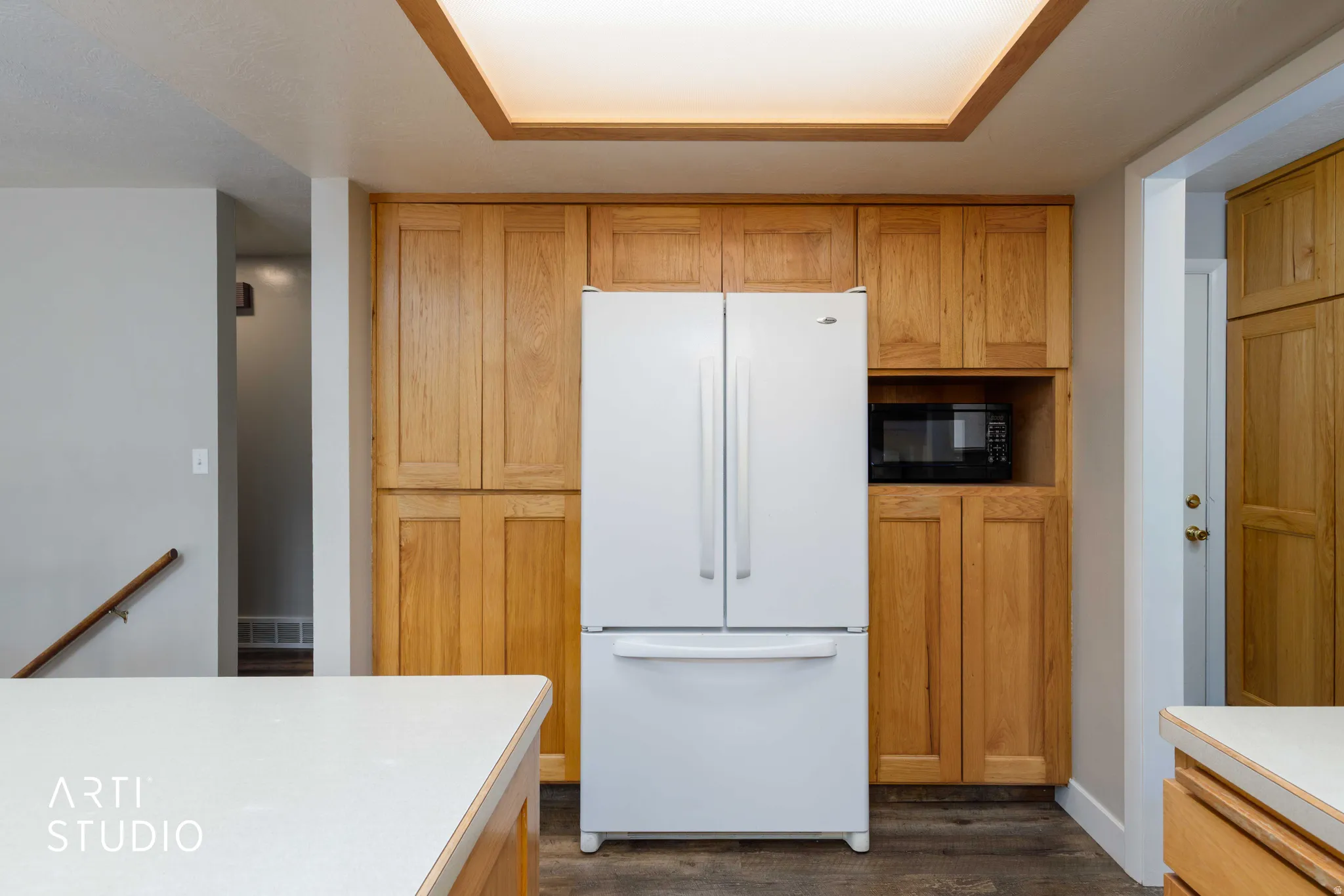 Integrated refrigerator and microwave within custom wood cabinetry, featuring a large ceiling light panel, and wood-finish flooring