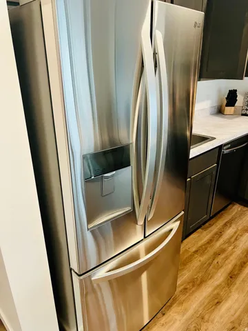 Stainless steel French door refrigerator with integrated dispenser, dark wood-finish cabinetry, light-toned countertop, and wood-finish flooring