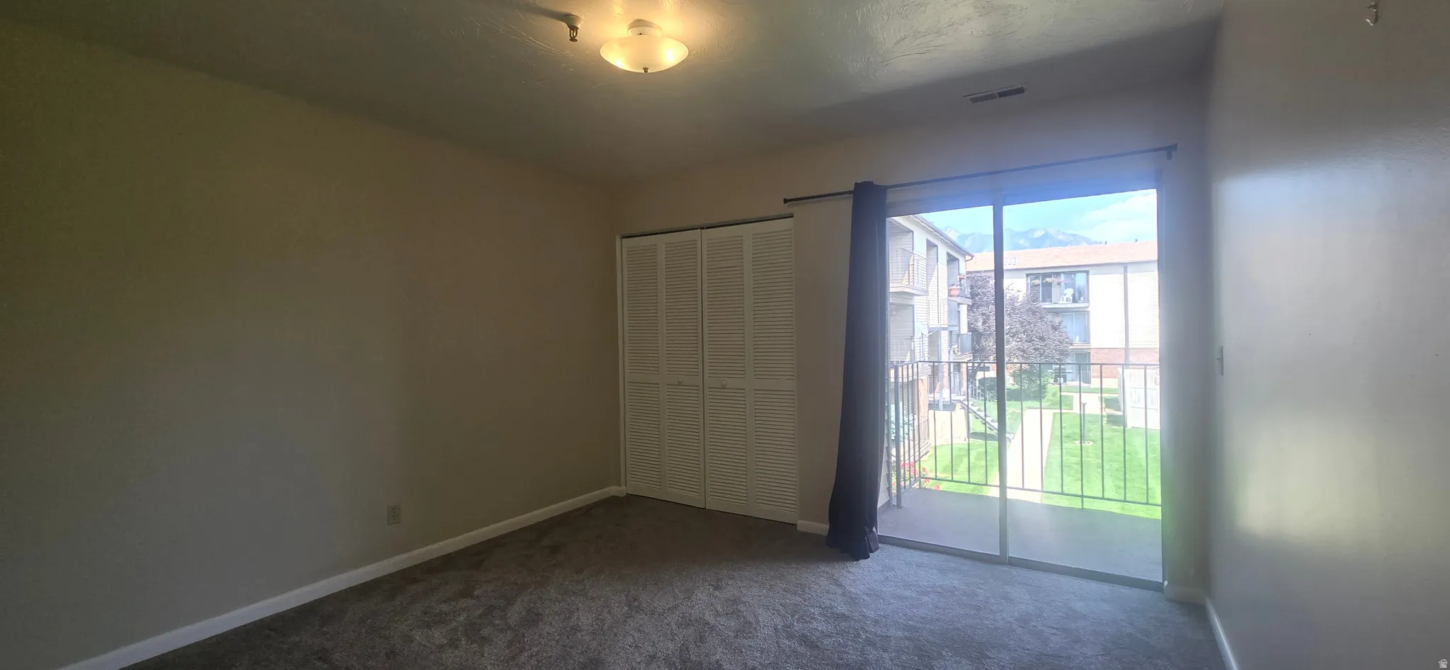 Neutral wall paint, ceiling-mounted light fixture, sliding glass door with balcony access, white louvered bifold closet doors, and gray carpet flooring