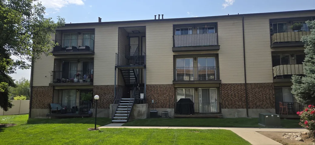 Three-story multi-unit building featuring exterior balconies, tan horizontal siding, and a partial stone-look facade