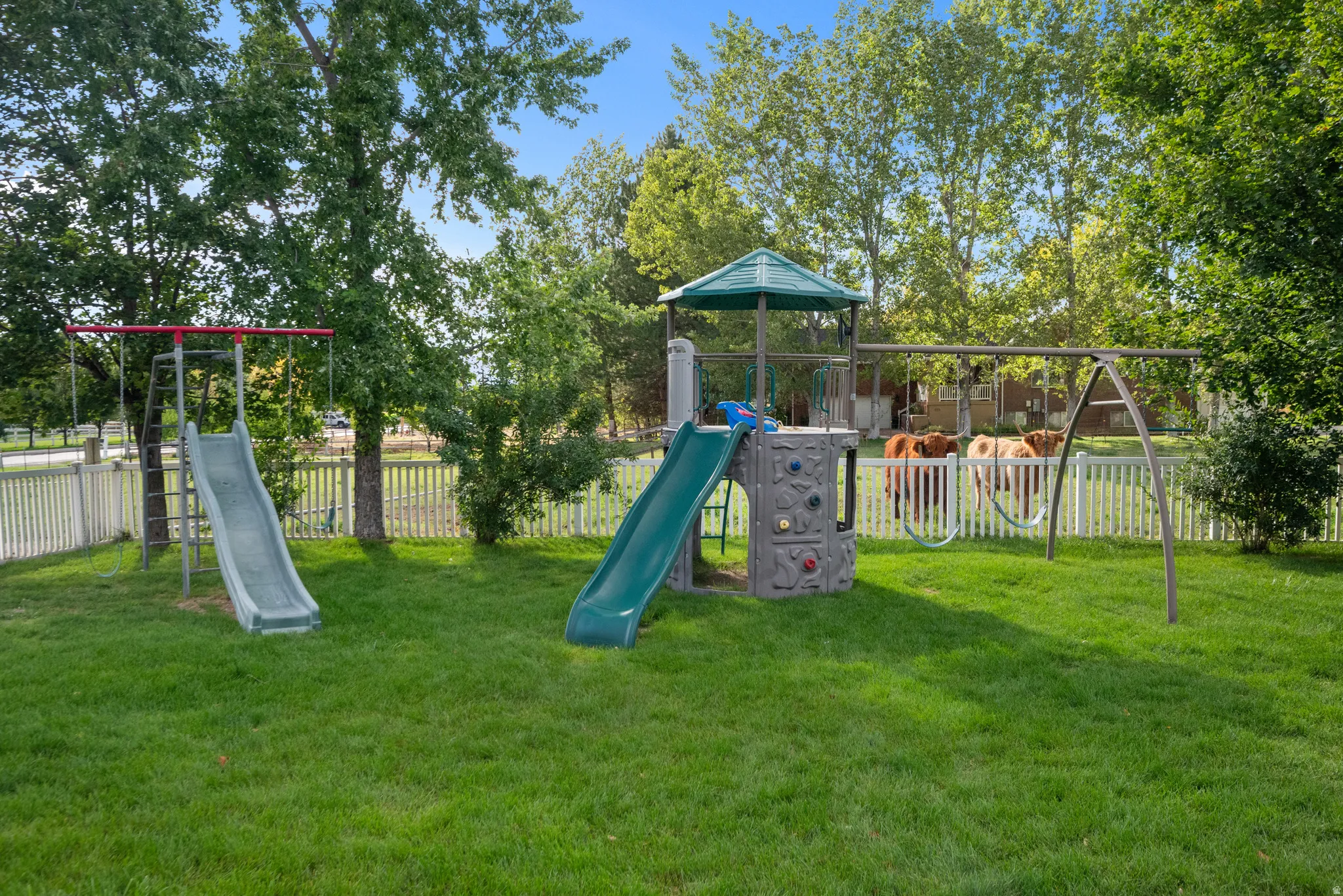 Expansive grassy yard featuring a white picket fence, mature trees, and a play structure with slides and swings