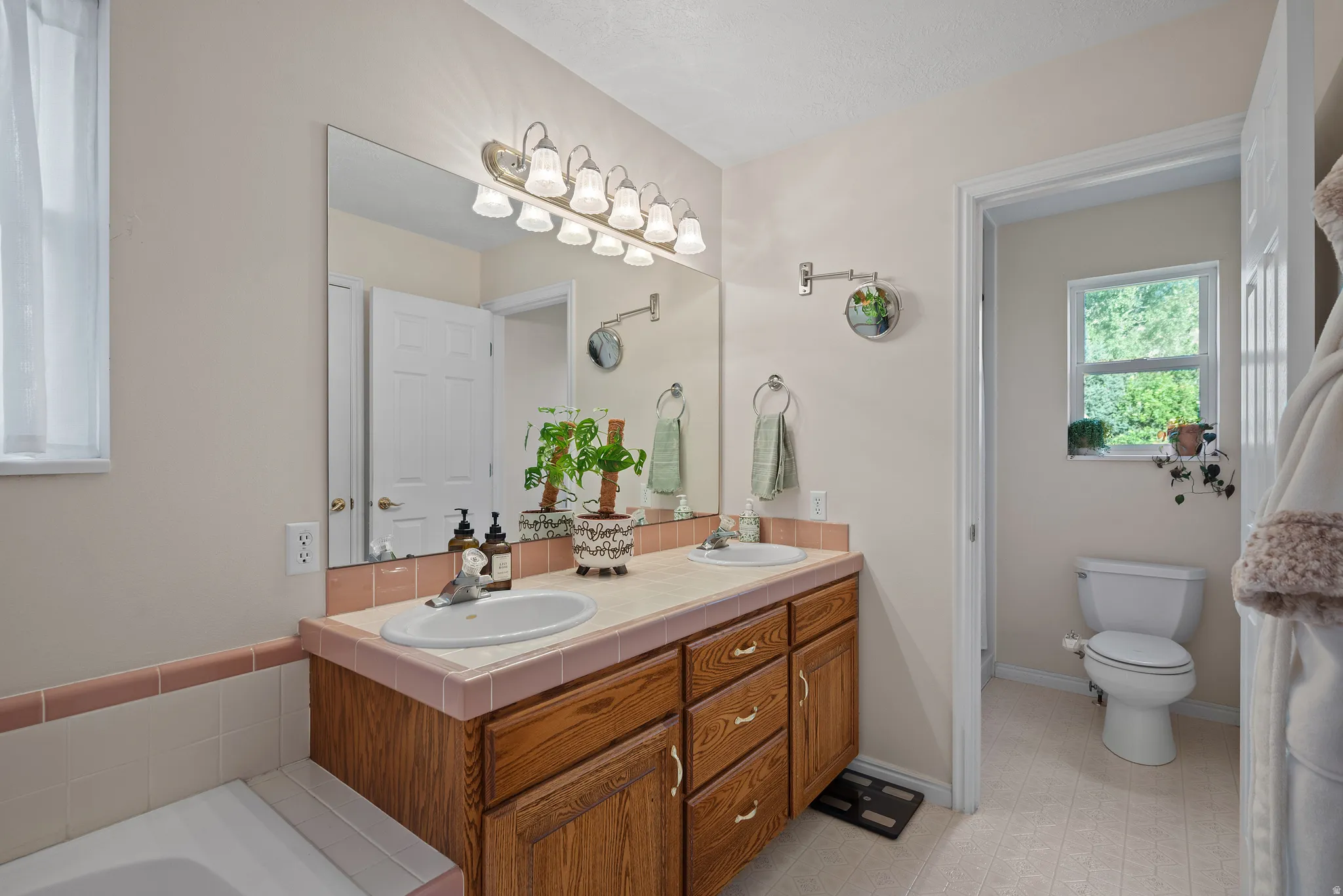 Dual basin vanity with wood-finish cabinetry, tile countertop, and a large mirror with integrated lighting