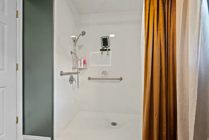 White shower stall featuring a built-in shelf, grab bars, and a handheld showerhead