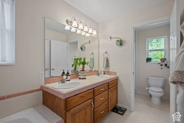 Dual basin vanity with wood-finish cabinetry, tile countertop, and a large mirror with integrated lighting