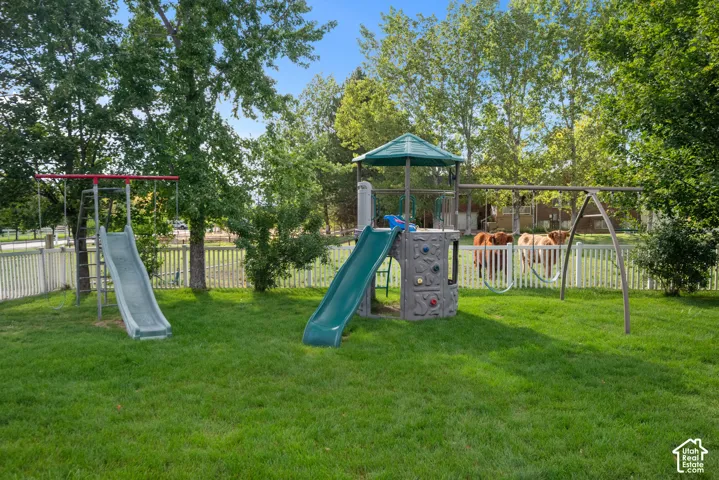 Expansive grassy yard featuring a white picket fence, mature trees, and a play structure with slides and swings