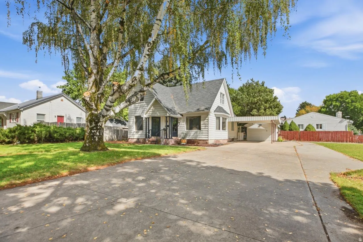 White siding exterior with a dark shingle roof, a paved driveway, a carport, and a mature deciduous tree