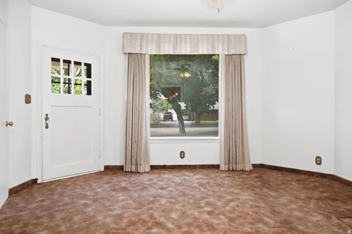 White paneled door with glass inserts, bay window with neutral drapes, ceiling fan, and brown carpeting- has its own entrance by front door