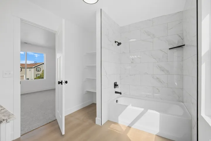 White soaking tub with matte black fixtures, marble-patterned tile surround, built-in shelving, and light wood-finish flooring
