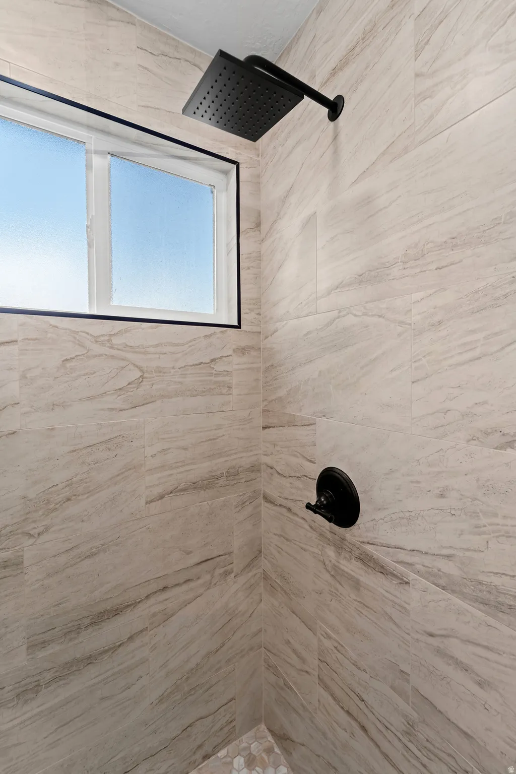Recessed window with black trim, matte black rainfall showerhead, matte black shower control, large format tile walls, and mosaic tile flooring
