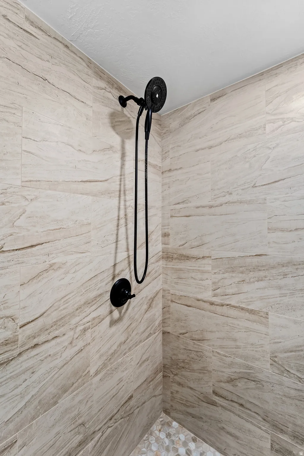 Shower enclosure featuring large format stone-look tile walls, matte black shower fixtures, and a pebble-style floor