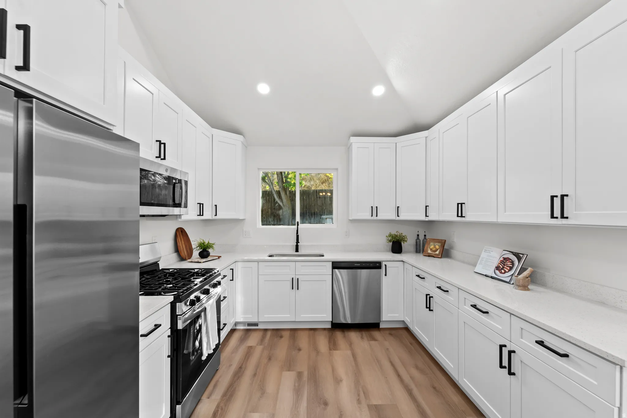 White shaker-style cabinetry with black hardware, stainless steel appliances, a white countertop, wood-finish flooring, and a window above the sink
