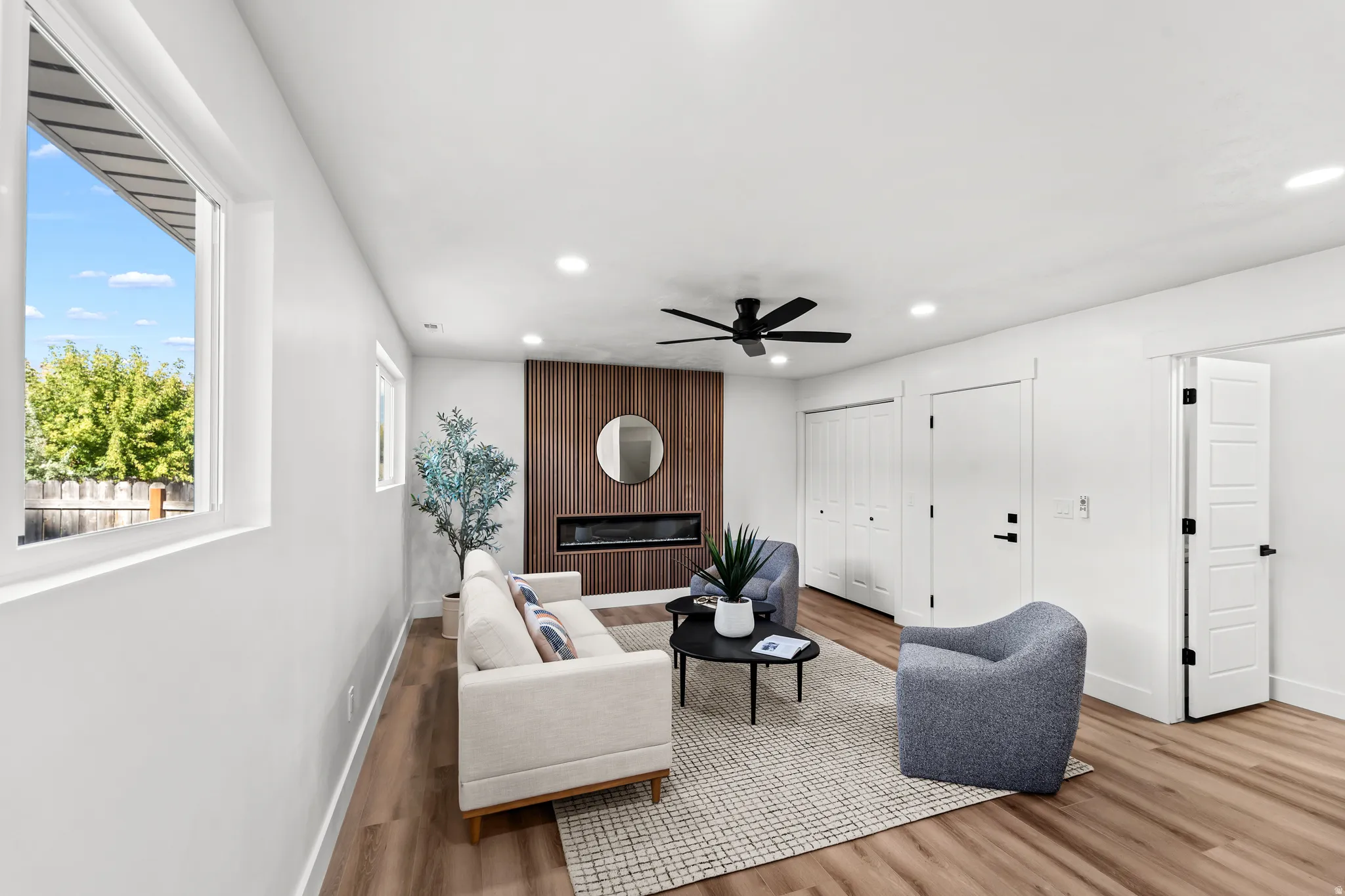 Spacious living area featuring wood-finish flooring, a contemporary ceiling fan, recessed lighting, a modern fireplace with a wood-paneled accent wall, and multiple windows