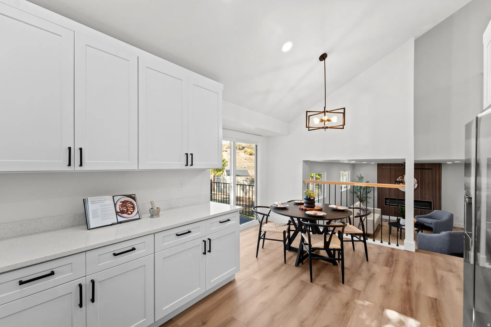 White shaker-style cabinetry with matte black hardware, light wood-finish flooring, a vaulted ceiling, a modern chandelier, and a sliding glass door