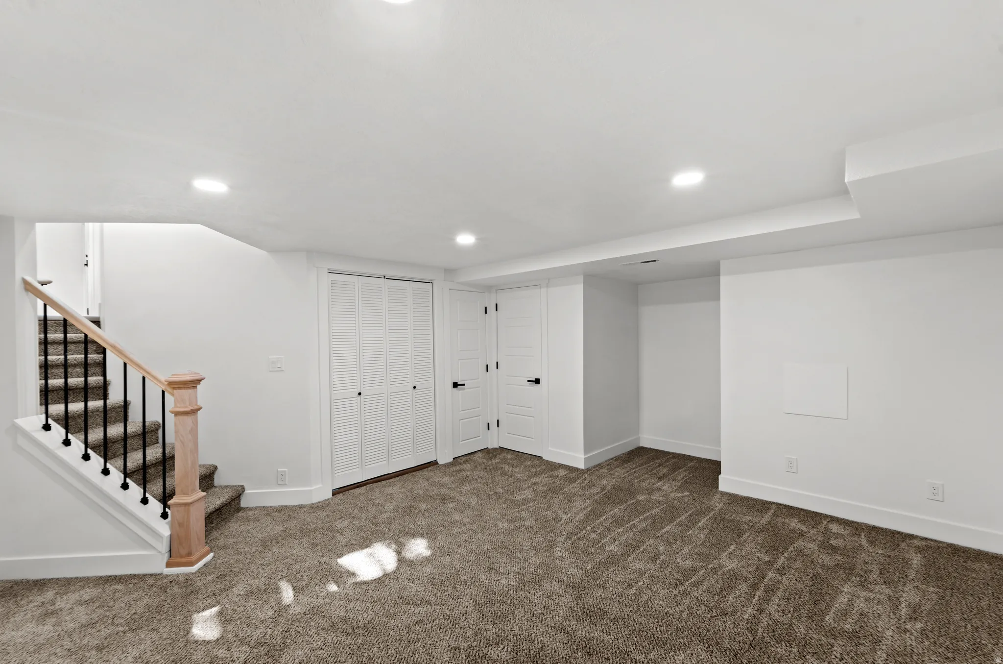 Recessed lighting illuminates this carpeted space featuring a staircase with wood newel post and wrought iron balusters