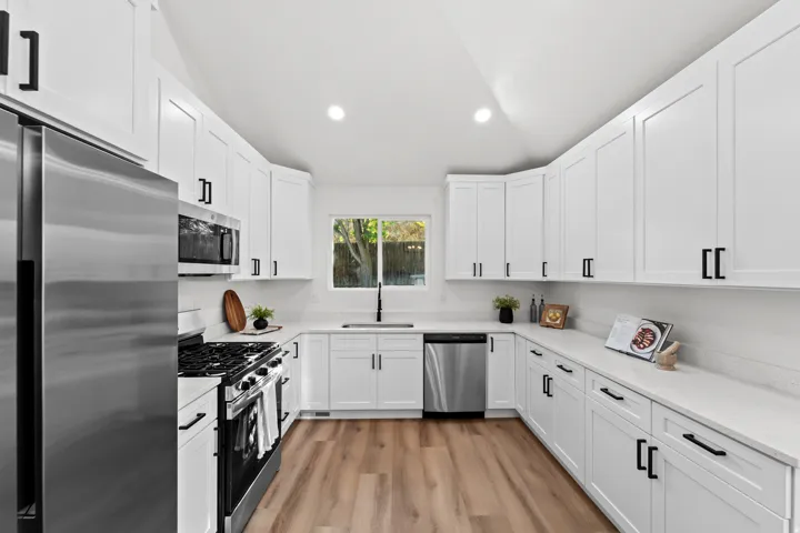White shaker-style cabinetry with black hardware, stainless steel appliances, a white countertop, wood-finish flooring, and a window above the sink