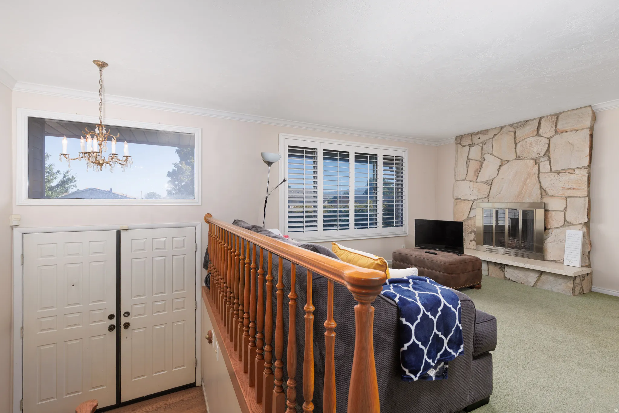 Stone-faced fireplace with a raised hearth, wood railing, recessed window with plantation shutters, decorative chandelier, and paneled double entry doors