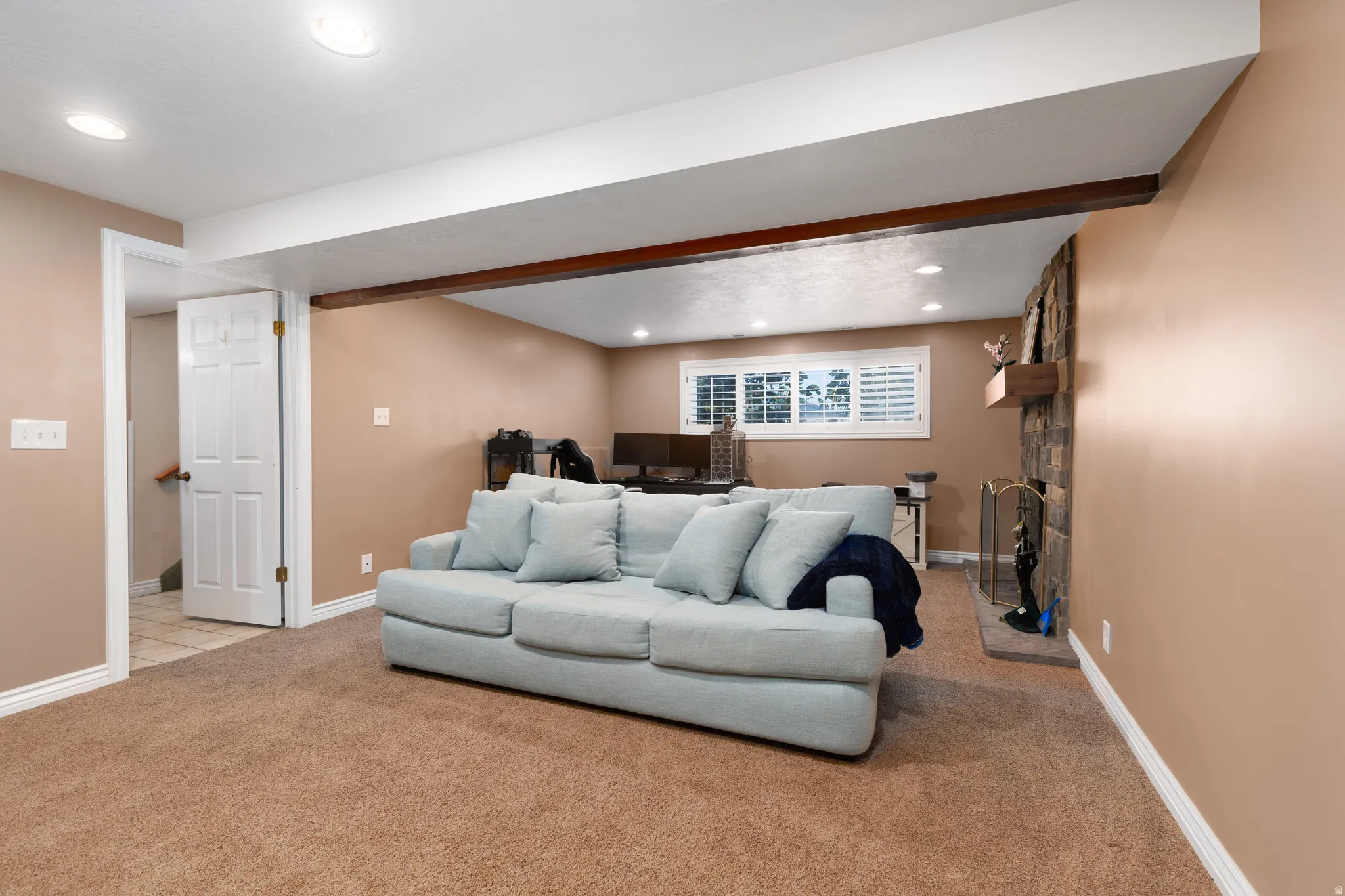 Carpeted living space featuring a stone-clad fireplace, recessed lighting, and a white plantation-shuttered window