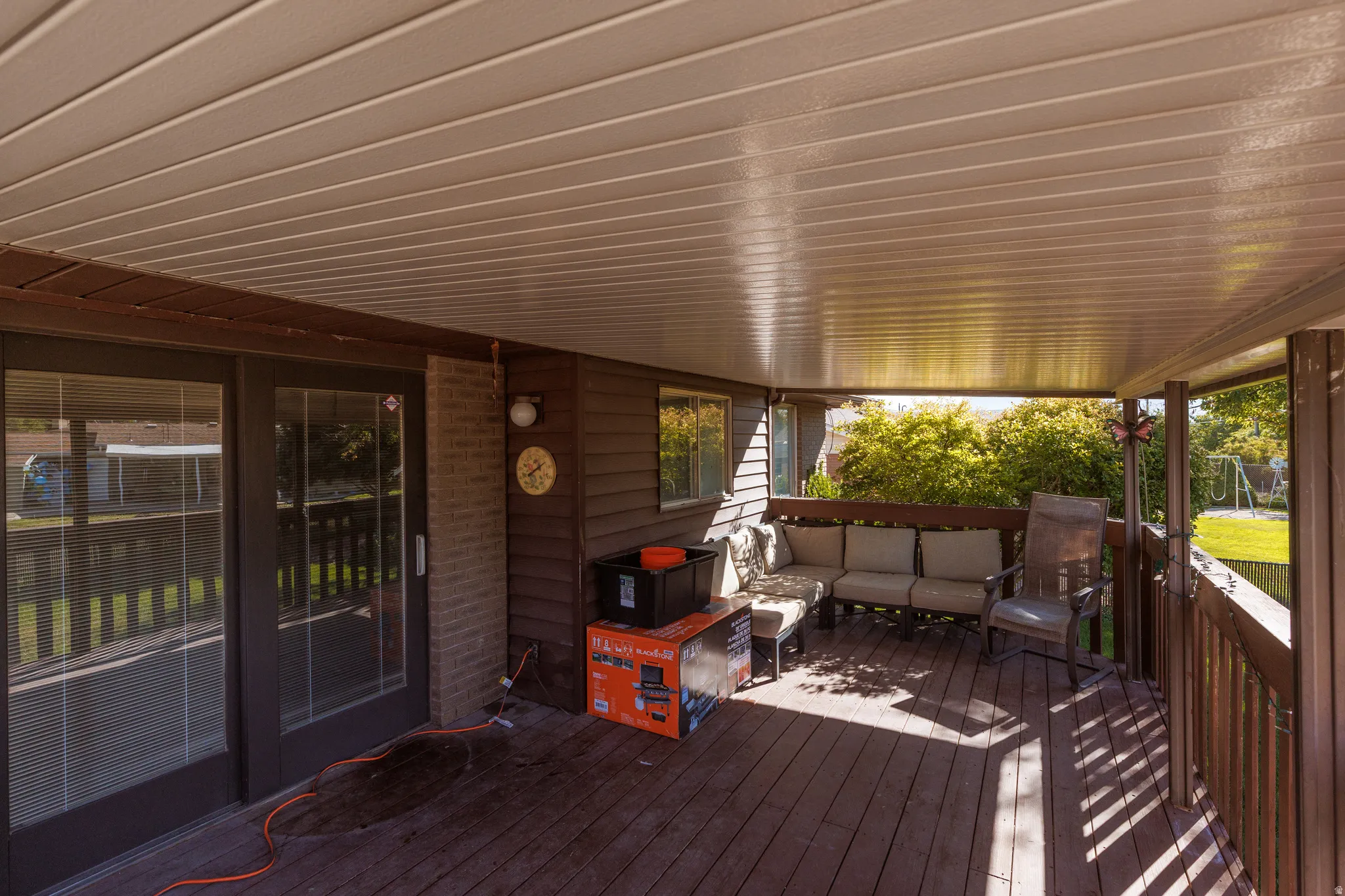 Covered deck with wood-finish flooring, featuring sliding glass doors, horizontal siding, and a partial brick wall