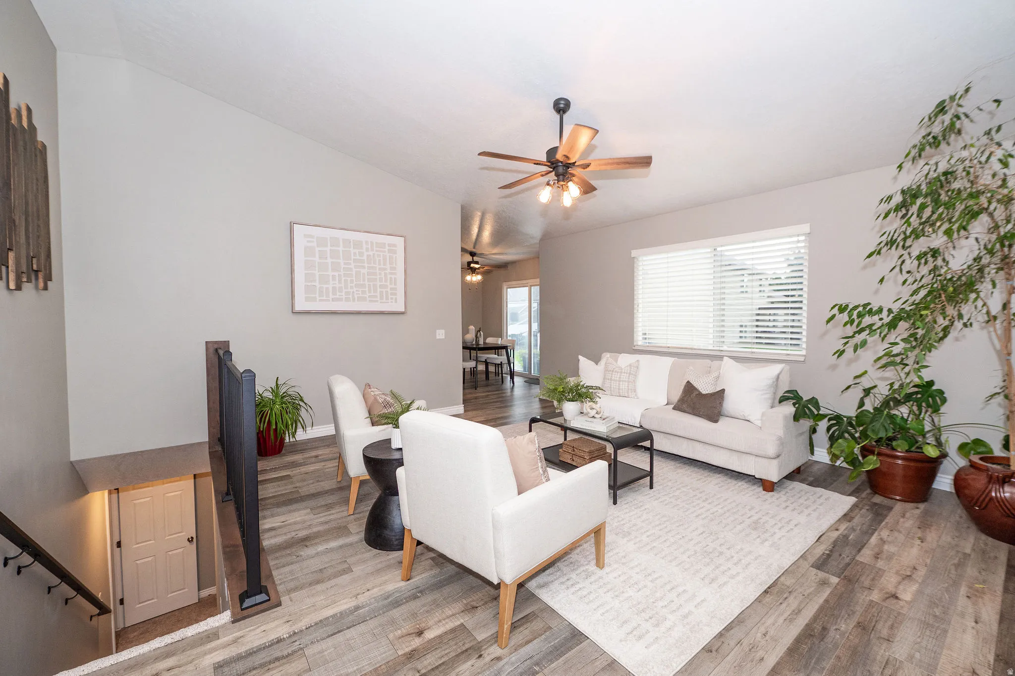 Vaulted ceiling living area with wood-finish flooring, a ceiling fan, and a large window with blinds