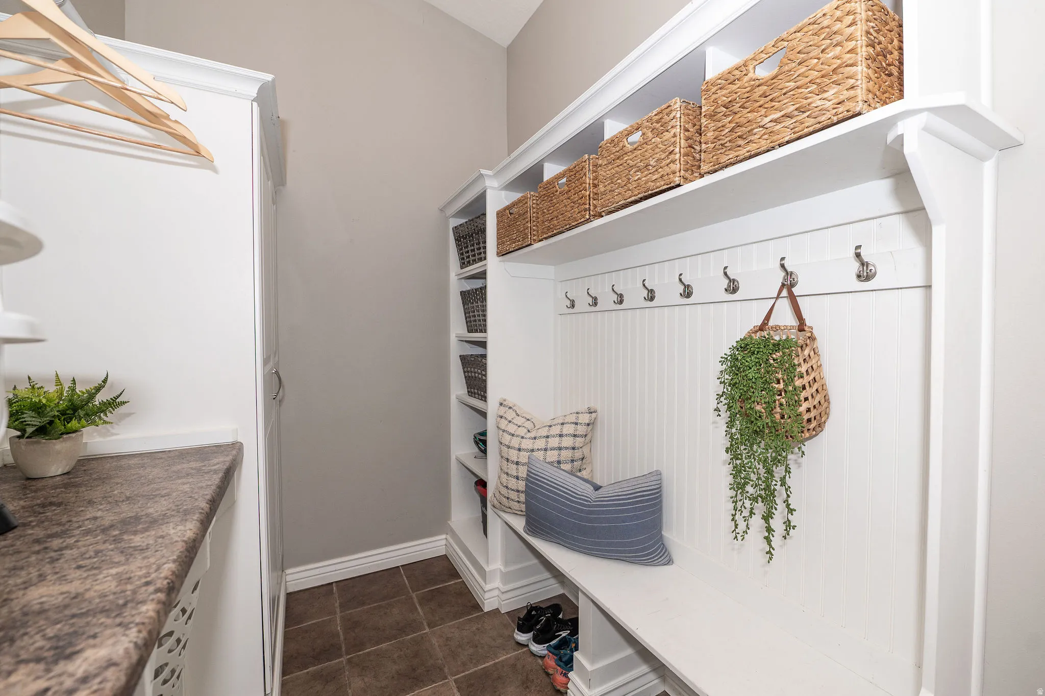 Built-in mudroom bench with coat hooks, overhead shelving, tiled flooring, and a stone-finish countertop
