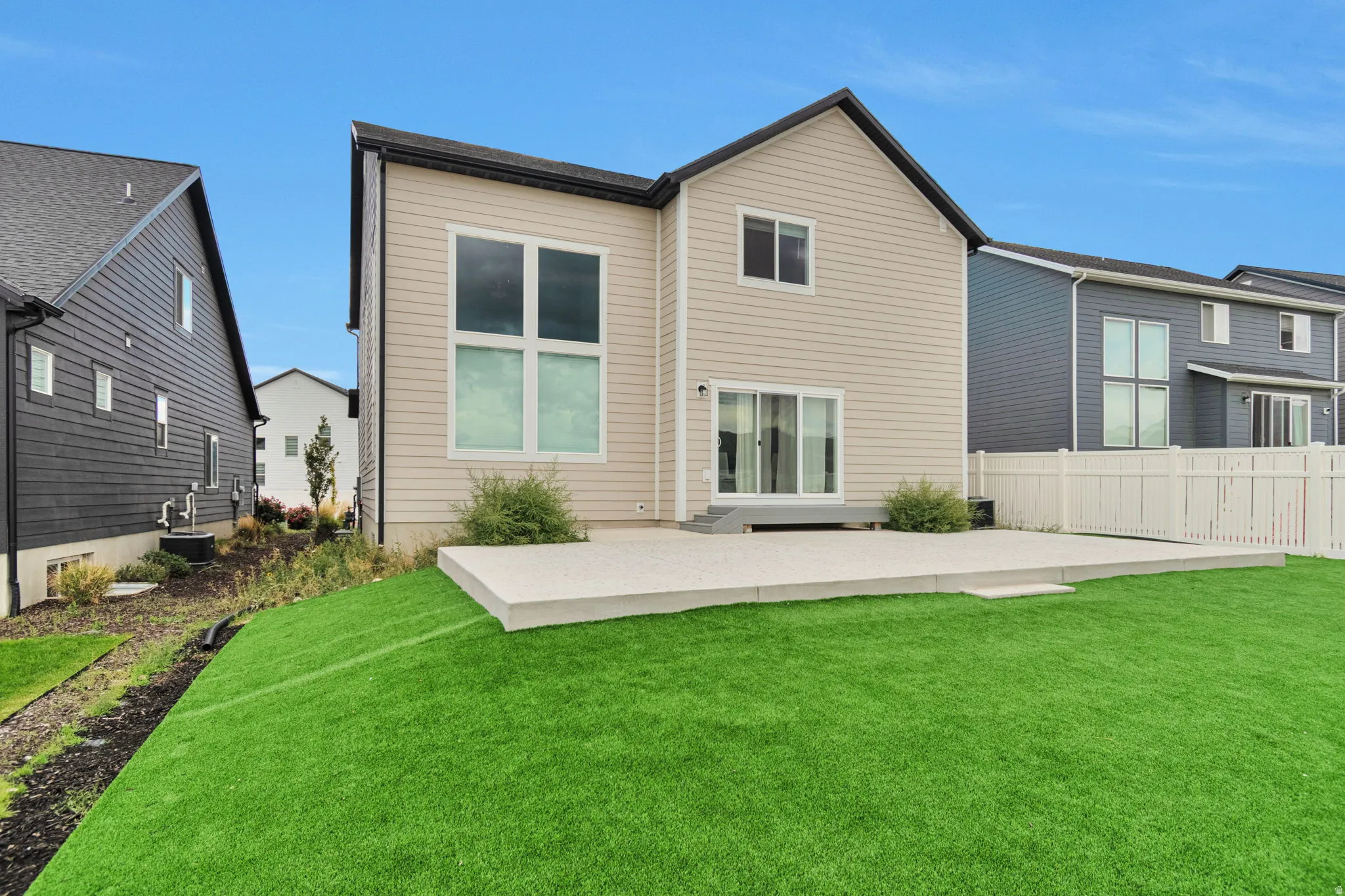 Expansive artificial turf lawn with a large concrete patio, white vinyl fencing, and sliding glass doors