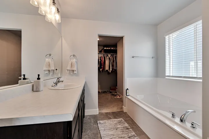 Spacious bathroom with a built-in soaking tub, vanity with an integrated sink, broad mirror, and a window with horizontal blinds