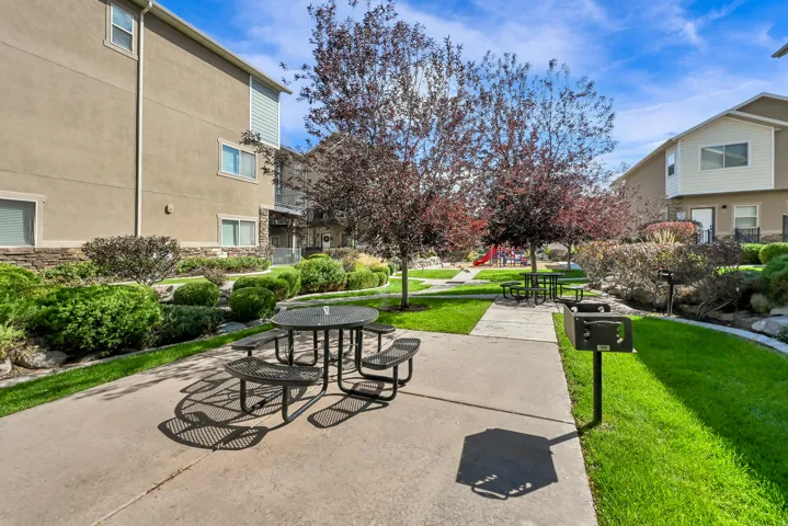 Manicured common area featuring mature trees, landscaped shrubbery, a concrete patio with a picnic table, and a built-in barbecue grill