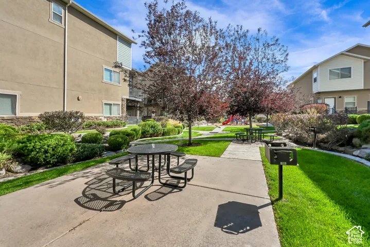 Manicured common area featuring mature trees, landscaped shrubbery, a concrete patio with a picnic table, and a built-in barbecue grill