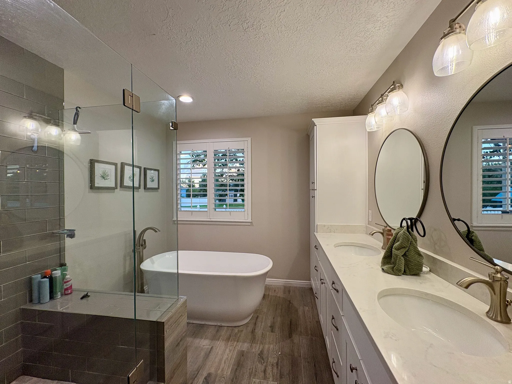 Freestanding soaking tub, glass-enclosed shower with tiled bench, dual vanity with undermount sinks and light countertops, wood-finish flooring, and window with plantation shutters