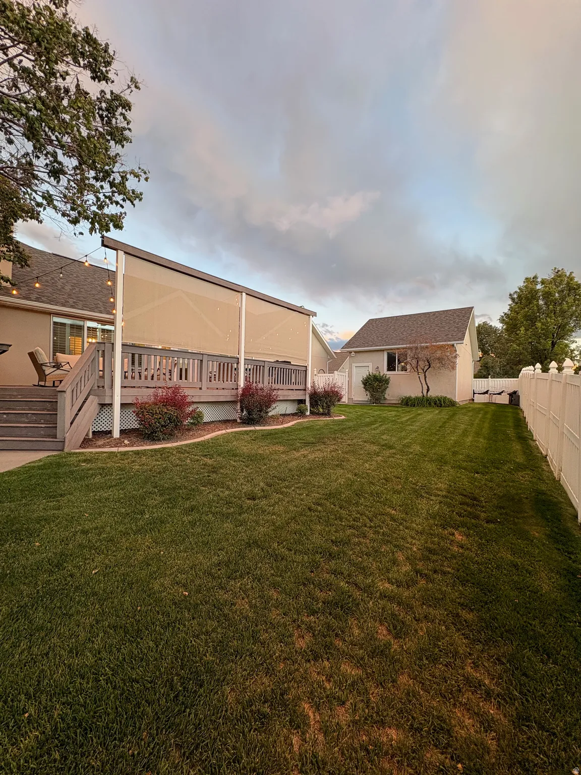 Expansive lawn area with a deck featuring a retractable shade system, a white privacy fence, and an accessory building