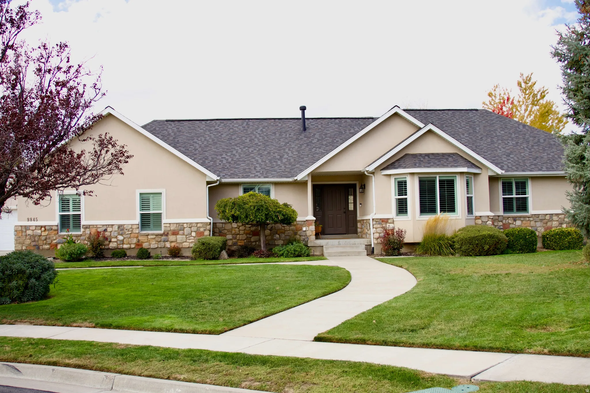 Stucco exterior with stone accents, a dark shingle roof, a curving concrete walkway, and established landscaping