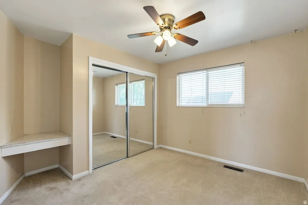 Built-in corner desk, mirrored closet doors, dual windows with horizontal blinds, and a ceiling fan with integrated lighting