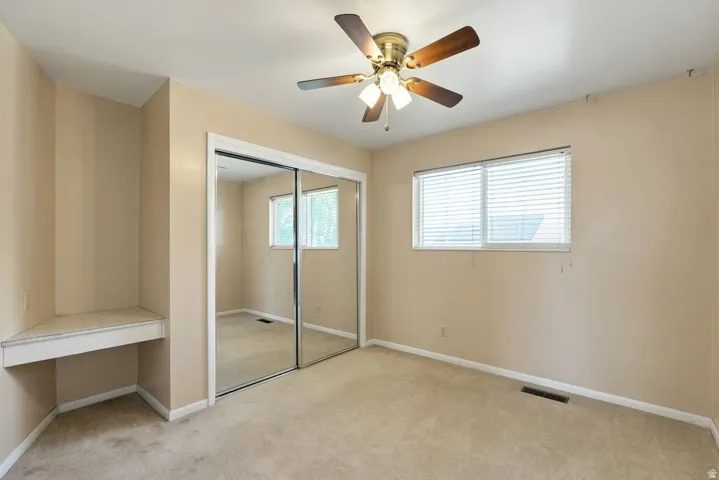 Built-in corner desk, mirrored closet doors, dual windows with horizontal blinds, and a ceiling fan with integrated lighting