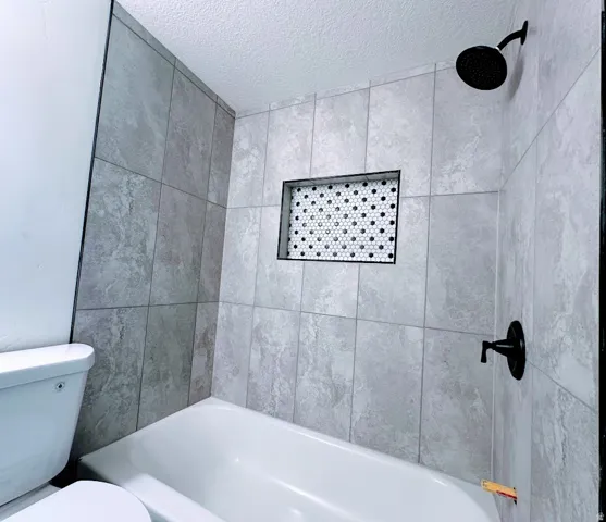 Bathtub with light gray tile surround, matte black shower fixtures, and a recessed niche featuring hexagonal mosaic tile