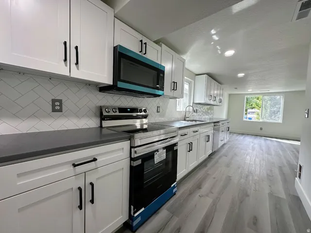 Sleek kitchen featuring white shaker cabinetry, dark countertops, herringbone tile backsplash, stainless steel appliances, and wood-finish flooring