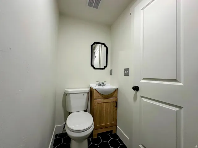 Black hexagonal floor tiling, wood-finish vanity with integrated sink, contemporary wall mirror, panel door, and standard toilet