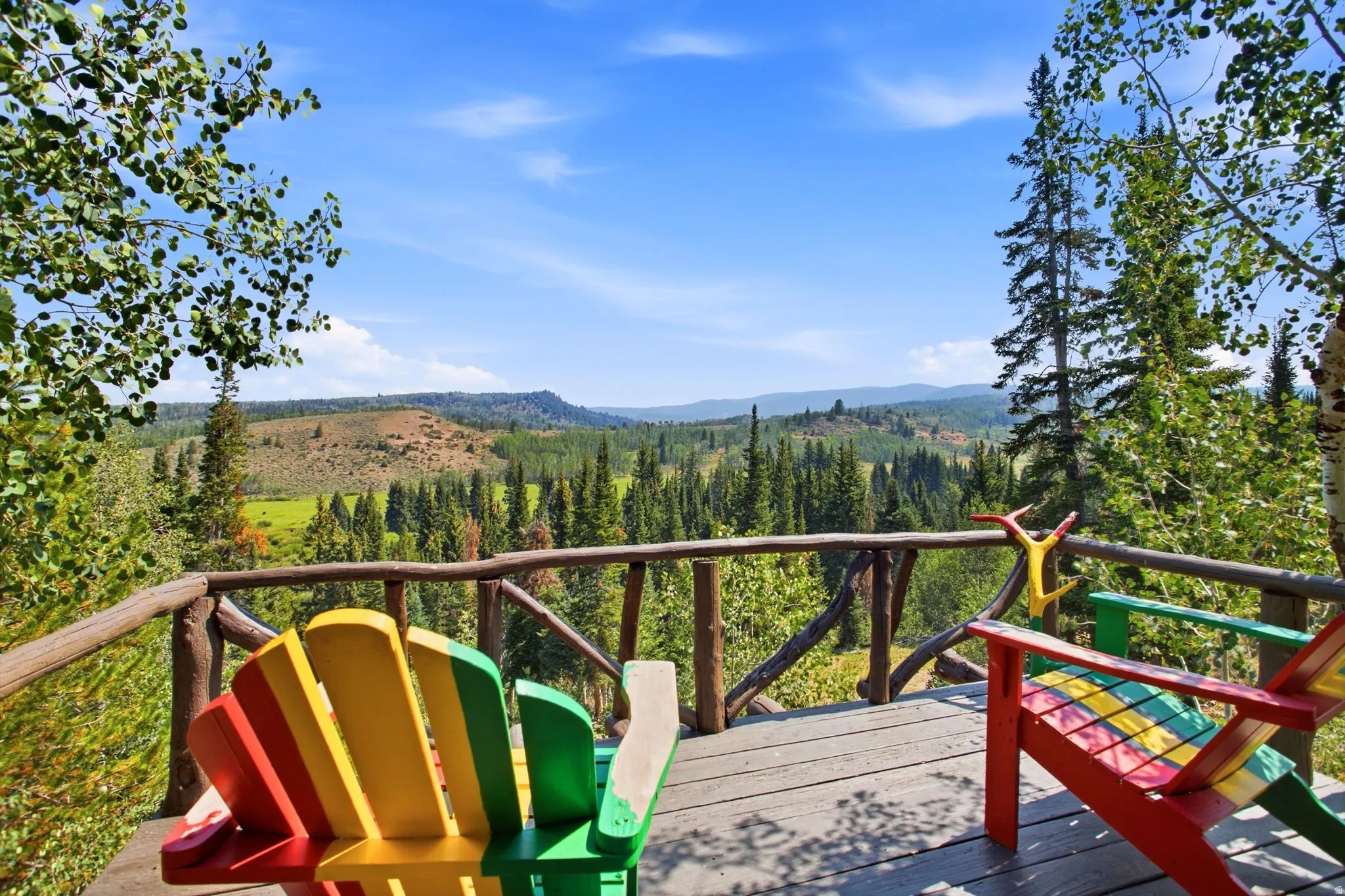 Expansive wood-plank deck with a rustic branch railing, surrounded by mature trees, overlooking a distant forested landscape