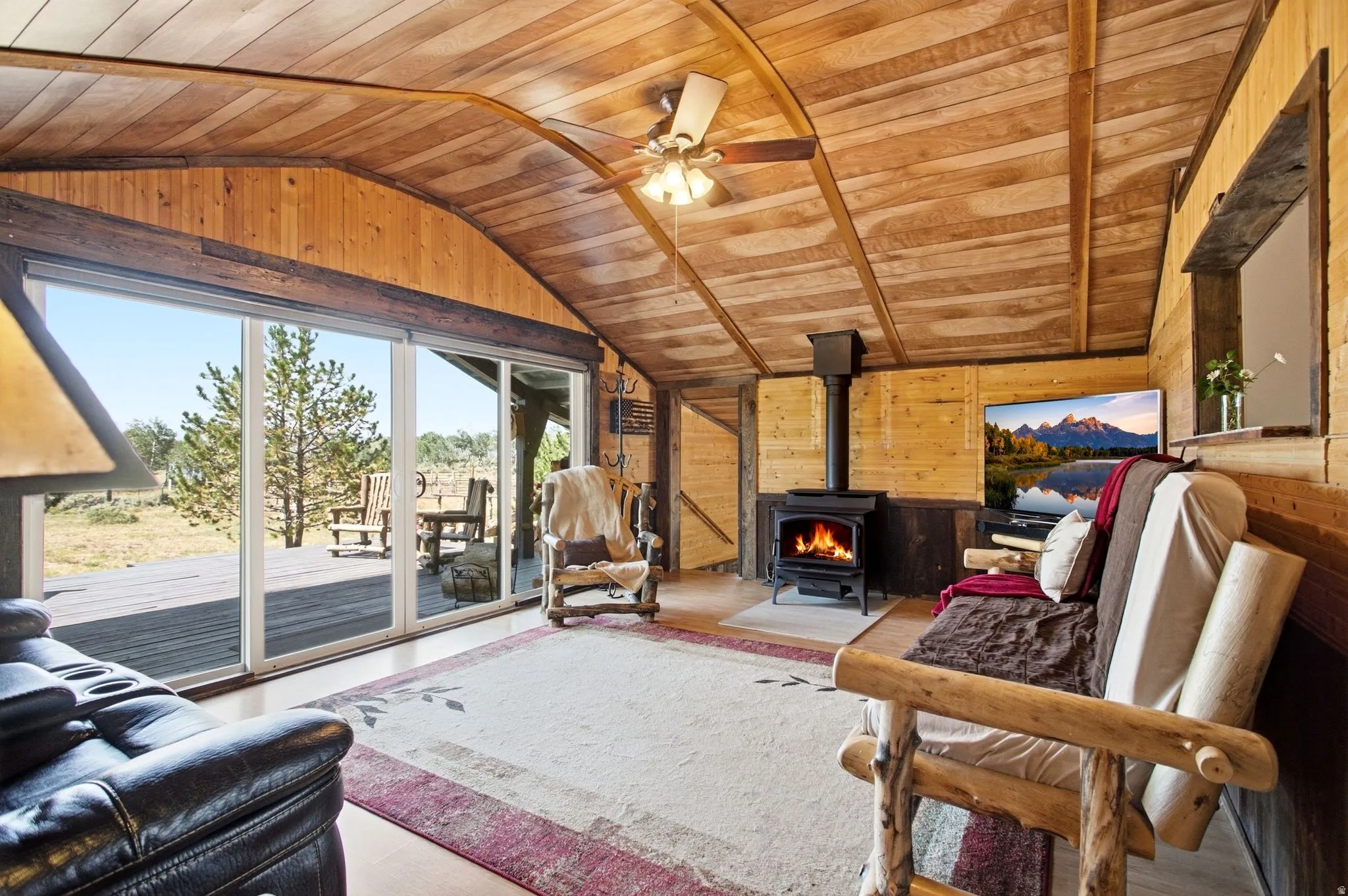 Vaulted wood-paneled ceilings, wood-finish walls, a freestanding wood-burning stove, and expansive sliding glass doors opening to a wood-plank deck