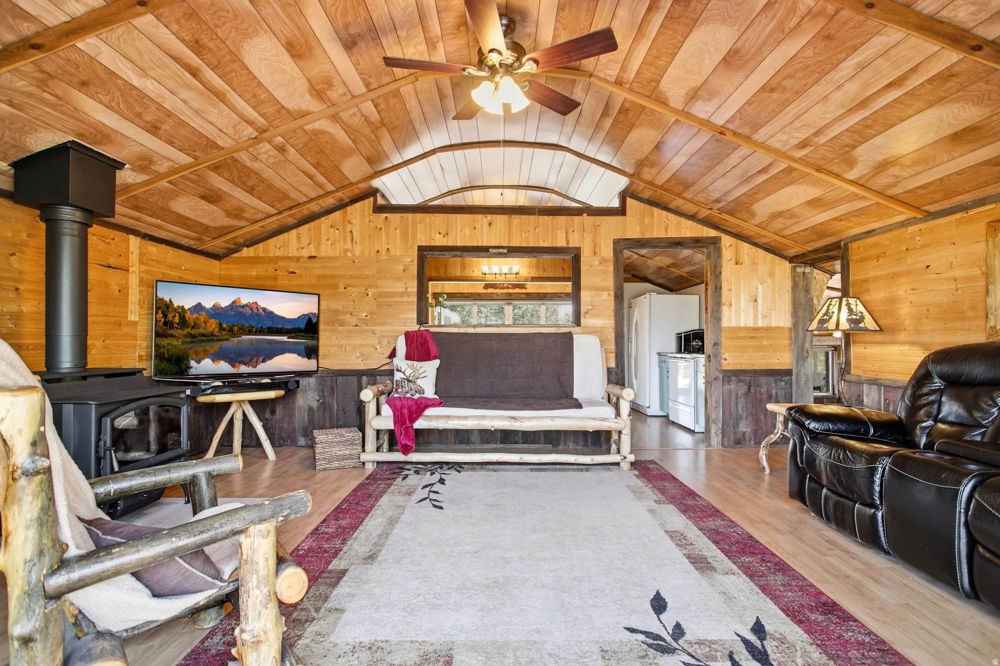 Vaulted wood-paneled ceiling with a ceiling fan, wood-finish flooring, wood-paneled walls, and a black wood-burning stove