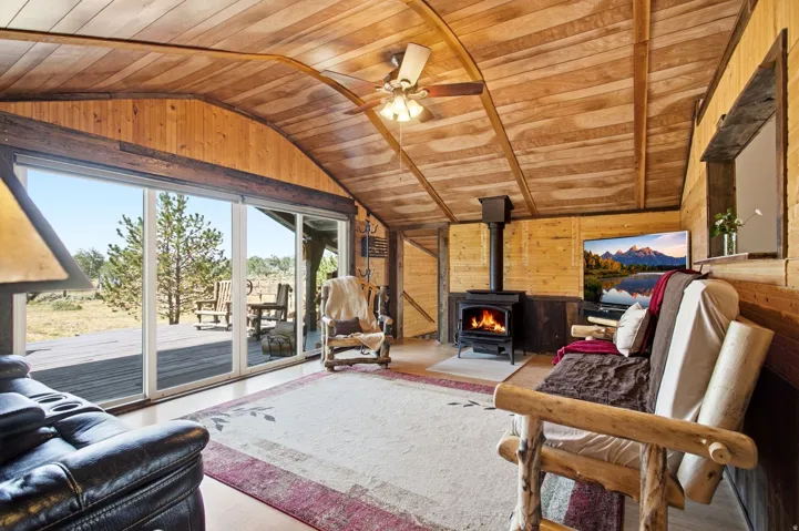 Vaulted wood-paneled ceilings, wood-finish walls, a freestanding wood-burning stove, and expansive sliding glass doors opening to a wood-plank deck