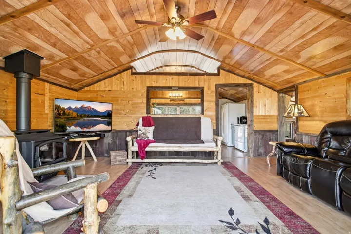 Vaulted wood-paneled ceiling with a ceiling fan, wood-finish flooring, wood-paneled walls, and a black wood-burning stove