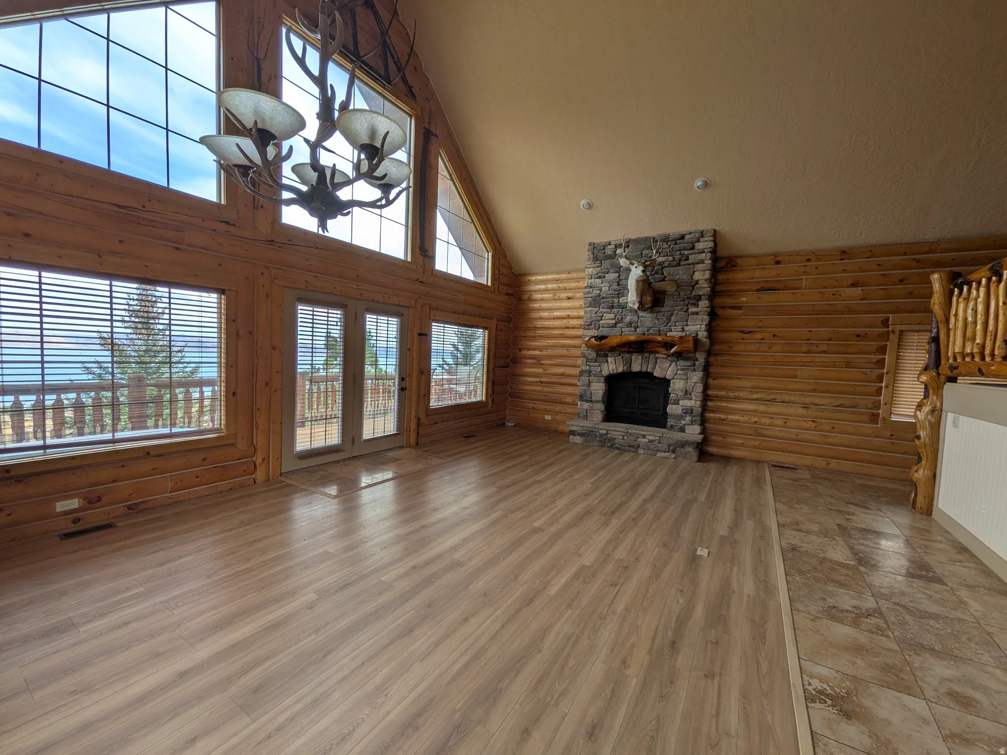 Vaulted ceiling with a decorative chandelier, expansive windows, stone-clad fireplace with a wood mantel, wood-finish flooring, and natural wood log walls