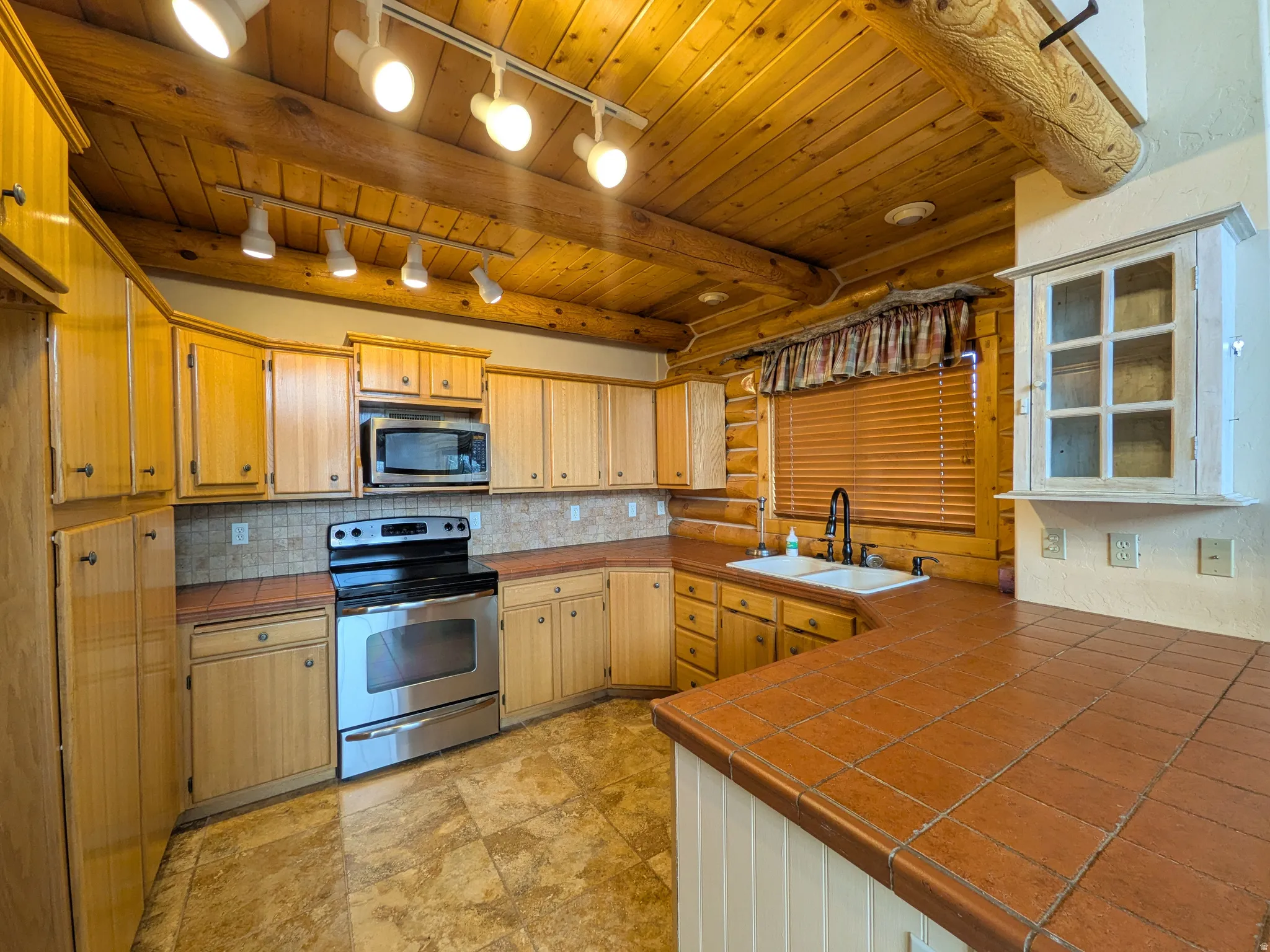 Wood-paneled vaulted ceiling with exposed beams, track lighting, light wood cabinetry, stainless steel appliances, and tiled countertops
