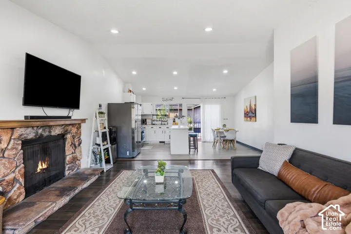 Open-concept living space featuring a stone-clad fireplace, dark wood-finish flooring, recessed lighting, and a white kitchen with an island