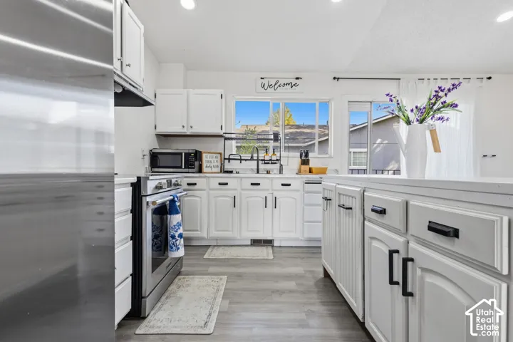 White cabinetry with matte black hardware, stainless steel appliances, recessed ceiling lights, and wood-finish flooring