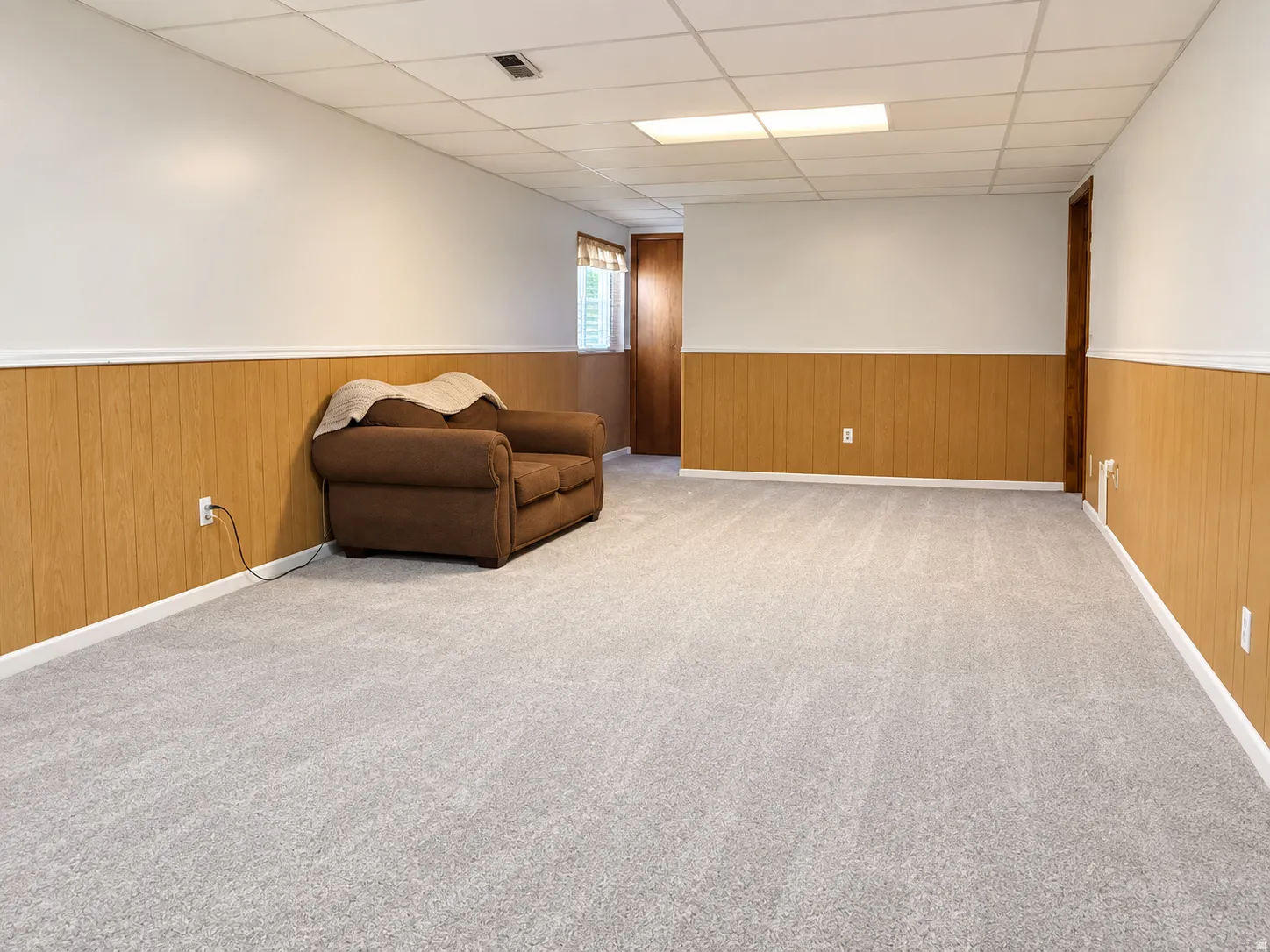 Spacious room featuring light grey carpet, wood-finish wainscoting, white painted walls, and a drop ceiling with integrated lighting